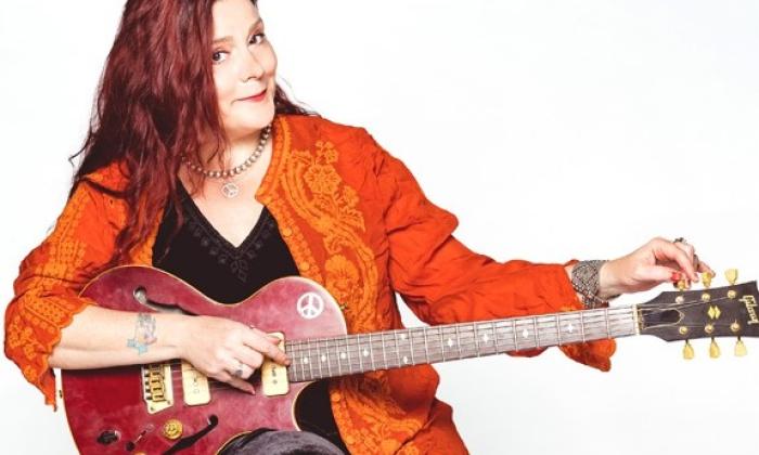 Carolyn Wonderland smiles while holding a guitar in front of a white backdrop.