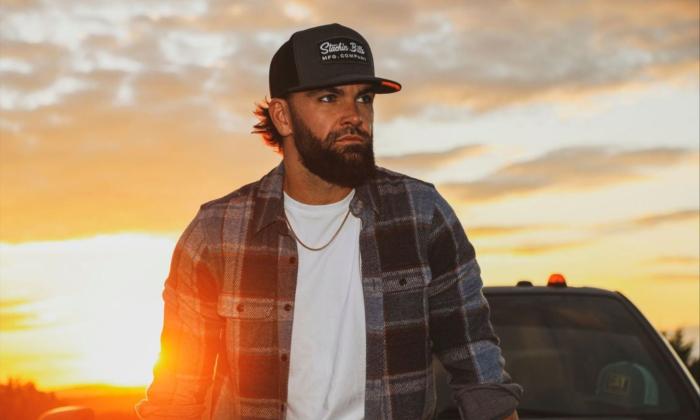 Dylan Scott dons a cap as he poses in front of his truck against a sunset backdrop.