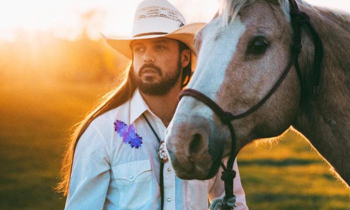 Ian Munsick stands beside a horse, with a scenic pasture stretching behind him. 