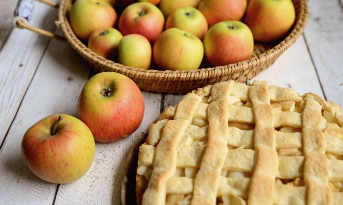 Apples sit on a table and in a basket, near a lattice-top apple pie