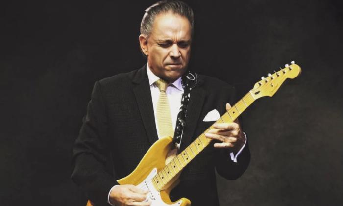 Jimmie Vaughan closes his eyes and strums his guitar in front of a black backdrop.