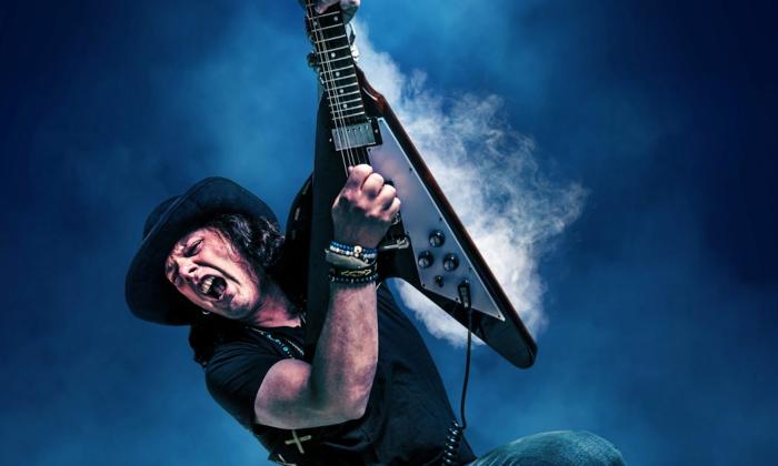 Anthony Gomes performs with his guitar raised high against a dark blue backdrop.