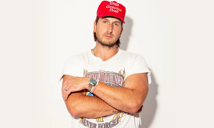 Russell Dickerson wears a bright red cap and folds his arms in front of a white backdrop.