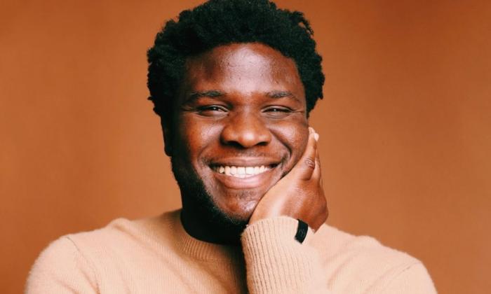 Tanael Joachim wears a light brown sweater and poses in front of a dark brown backdrop.