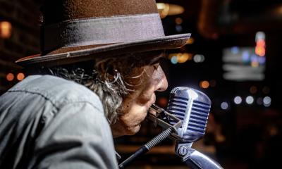 Bluesman Denny Blue in a fedora, playing harmonica into a microphone