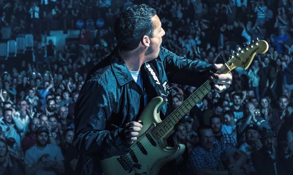 Performer Adam Sandler taken from behind on stage, holding a guitar and playing to a large, clapping crowd