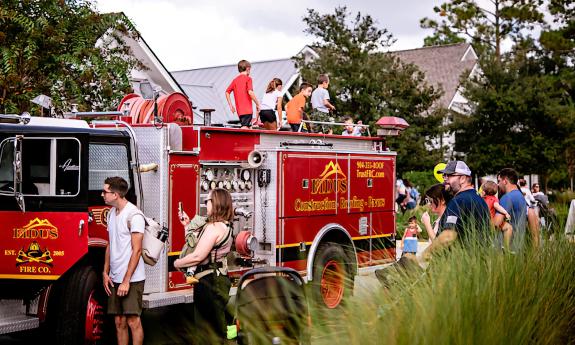 A firetruck near decorative grass with houses in the background and children climbing on the truck