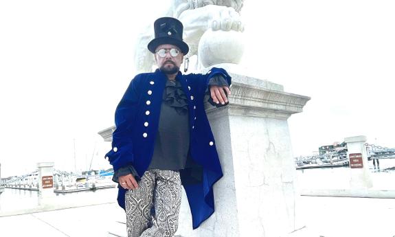 A man in dark Steampunk garb, leaning against the base of the lion, with the background pale