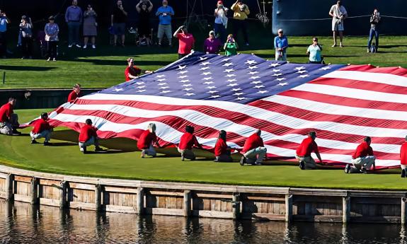 A large American Flag, held by people in red shirts on the banks of a pond