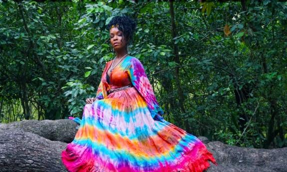 Kat Riggins wears a colorful dress and sits on the a large trunk in the forest