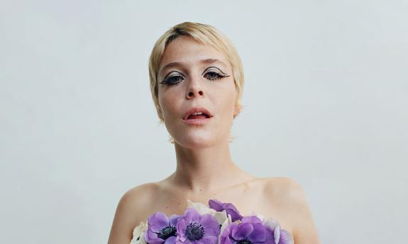 Maggie Rogers agaist a pale backdrop, wearing purple and white flowers