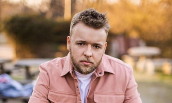 A young white man (singer Preston James) dressed in a pink jacket looks directly into the camera. Trees and cars in the background are blurred 