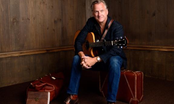 Ellis Paul smiles and poses with his guitar in front of a wooden wall. 