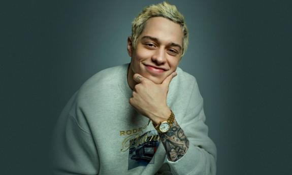 Comedian Pete Davidson smiles while clutching his chin. 