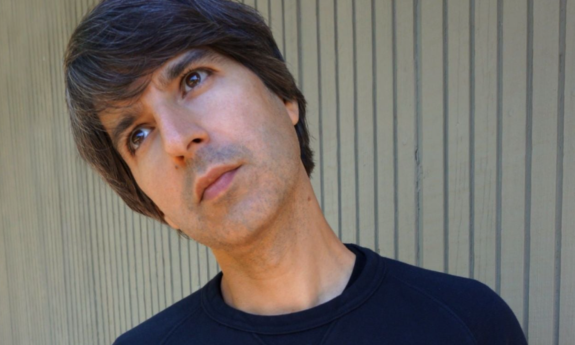 Demetri Martin flexes his neck and poses in front of a backdrop with stripes.