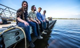 The Dirty Grass Players smile and pose on a dock while placing their feet close to water. 