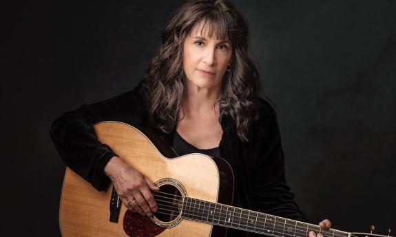 Karla Bonoff poses with her brown guitar in front of a black background. 