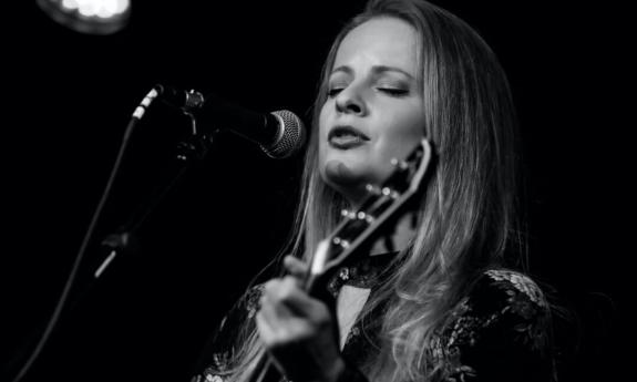 A black and white photograph of Alice Wallace singing on stage while strumming her guitar.