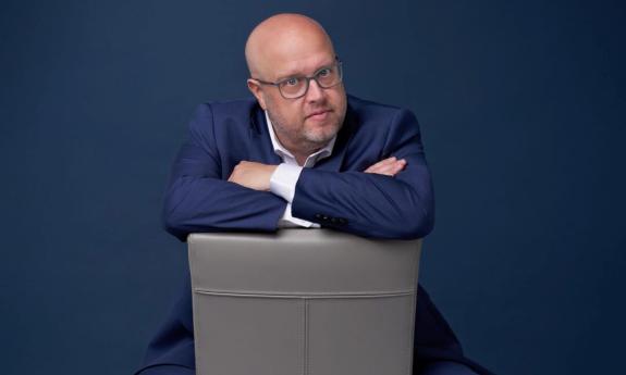 Comedian Danny Johnson poses in a blue blazer and glasses while sitting in a grey chair. 