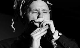 A black and white photograph of Jason Ricci playing the harmonica with his eyes closed. 