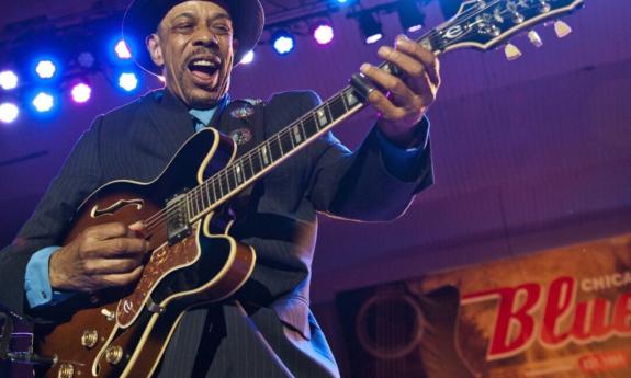 John Primer sings and strums his guitar on a well-lit stage. 