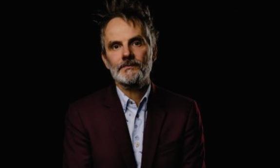 Mike Viola wears a red blazer and poses in front of a black background. 