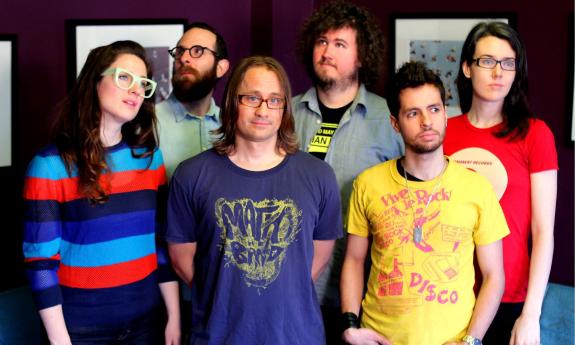 Bandmates from Wheatus pose in colorful shirts in front of a wall with framed pictures. 