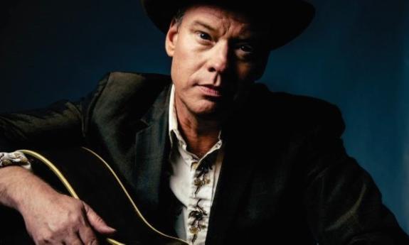 Shawn Mullins poses in dark clothing while strumming his guitar in front of a dark blue backdrop. 