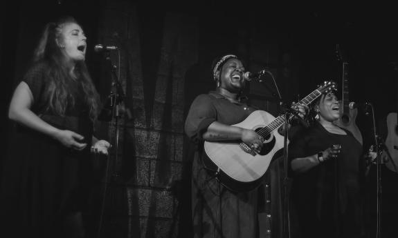 Three women singing on stage, one playing the guitar