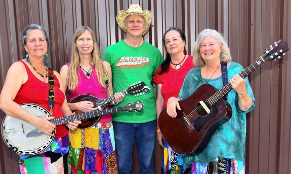 Five musicians, four women and one may, stand with string instruments in front of a barn wall