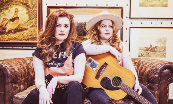 Cassie and Maggie pose side by side on a brown chesterfield couch in front of pictures set in a frame. 