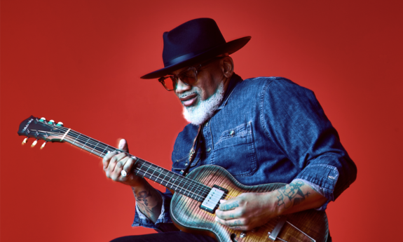 Toronzo Cannon wears black glasses while strumming his guitar in front of a red backdrop. 