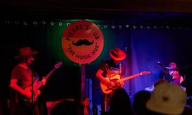 A band, wearing hats, on the stage of Pierre's Pub, in front of the pub's mustache sign