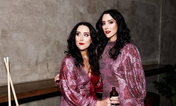 The Watson Twins standing in a lobby, wearing rose-colored dresses.