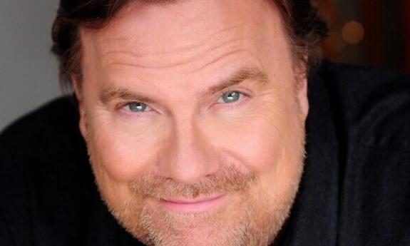 Kevin Farley smiles and poses in front of a grey background with bokeh. 