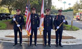 Four uniformed firefighters stand in formation, holding flags and ceremonial axes in honor of the fallen