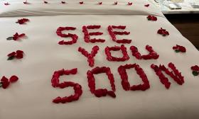 The words See You Soon, written in rose petals on a white bedspread