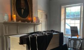 A plain wooden casket, near a window, being prepared for mourners