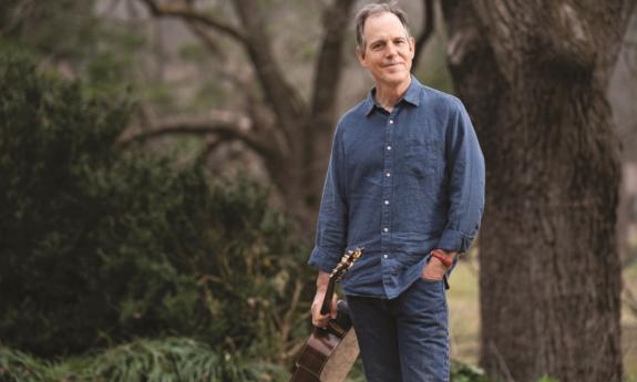 David Wilcox poses with his guitar in front of trees and shrubs. 