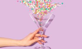 A woman's hand clutches a martini glass in front of a pink backdrop.