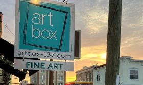 The sign for Art Box, white letters on a teal background, during sunset