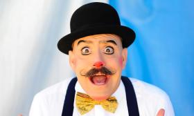 A clown with suspenders and a bowler and limited make-up beyond a red nose and mustache