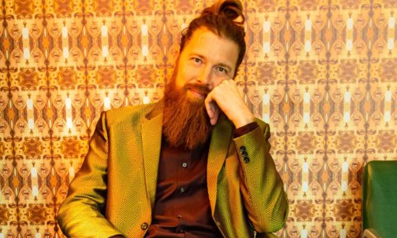 Shane Mauss sits in a chair and poses with a brown blazer and top in front of wallpaper with strange faces in the form of a pattern.