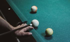 A male hand supports a billiard cue stick, lining up on the white ball close to the bumper