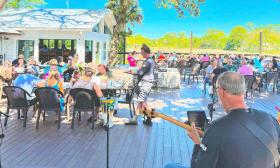 The deck of a seafood restaurant on a sunny afternoon, with a musician in the foreground