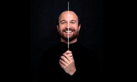 Composer and musician Juan Antonio Simarro in black, against a black backgroud, and holding a conductor's baton