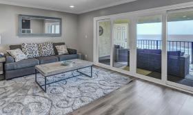 A living room with cream and blue has an expanse of glass open to the large furnished deck overlooking the ocean