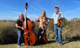 Members of a bluegrass and folk trio, standing in a field with their instruments under a blue sky