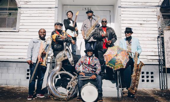 The seven members of the Dirty Dozen Brass Band, in front of an old, white building with their instruments