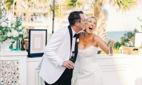 The bride and groom, both in white, laugh and hug near the celebration's bar